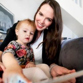 Mother and child sitting on a sofa and looking at a tablet. Child is sitting on mother's lap while the mother holds the tablet. The child is pointing to the tablet screen.